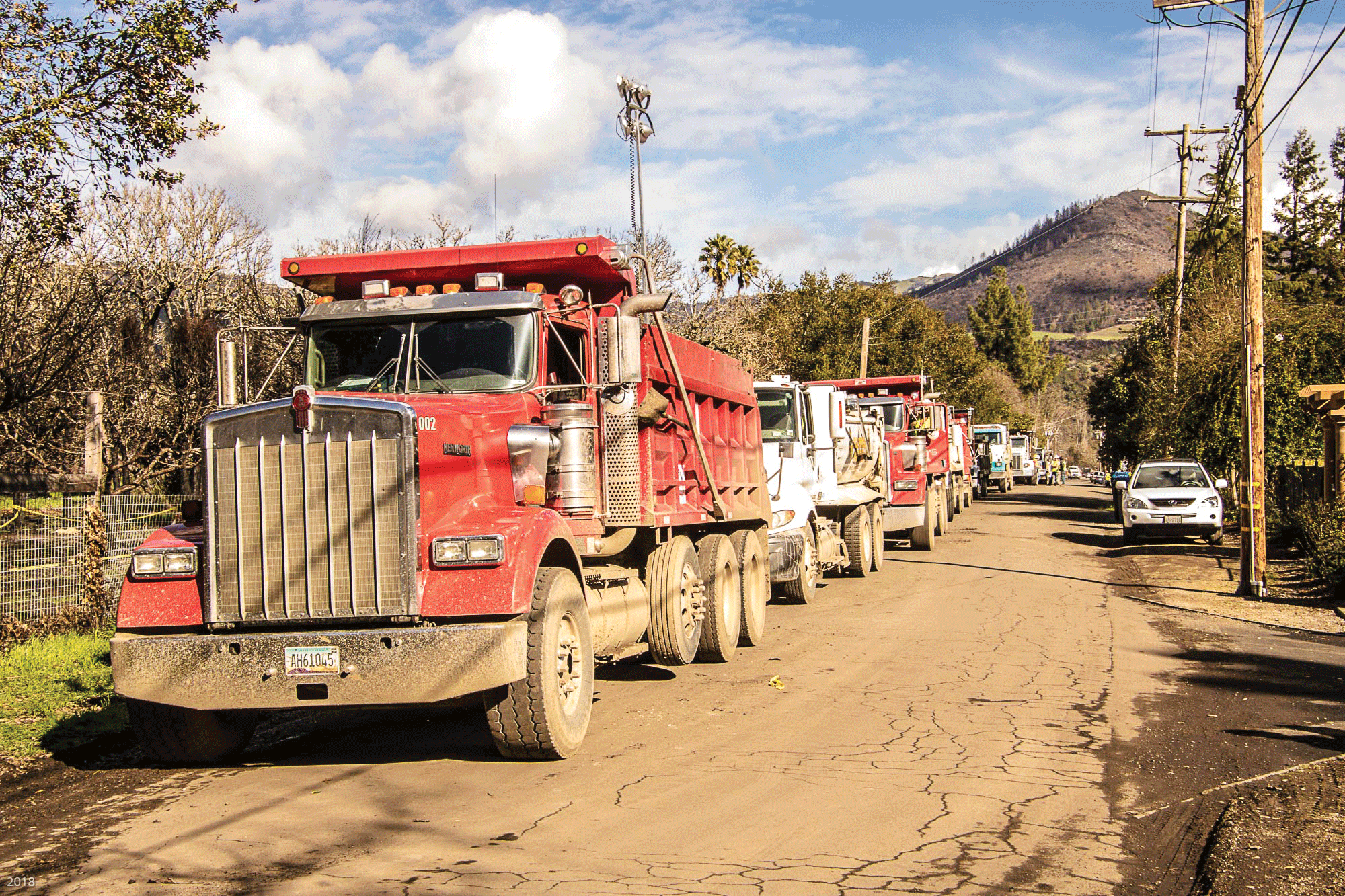 A row of dump trucks parked on the side of a narrow road, with a charred hillside in background