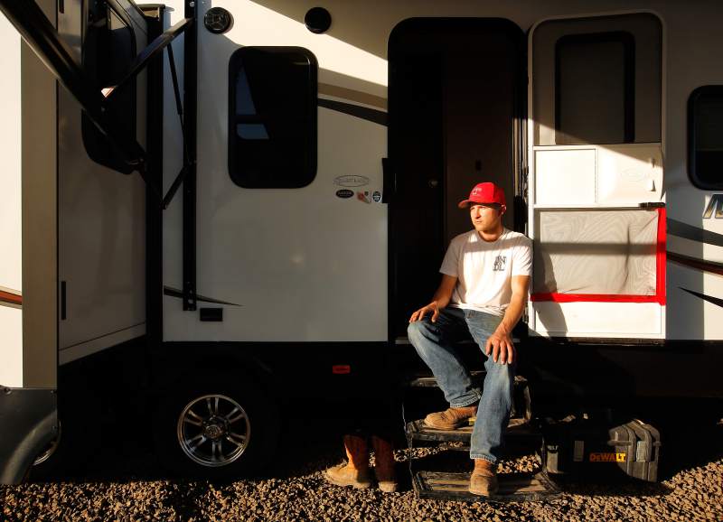 A person in a blue jeans, white t-shirt, leather work boots, and red baseball hat sits in the doorway of a trailer. Another pair of boots and a heavy duty toolbox rest on the ground nearby.