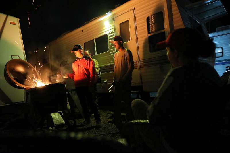 A group of people in t-shirts and sweaters stand around a raised fire pit near a camper trailer.